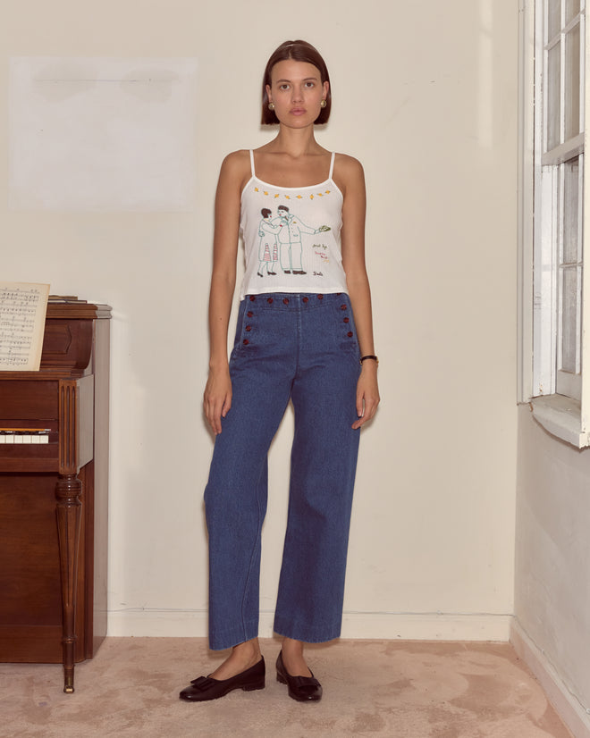 A woman wears the Companion Tank—a vintage motif embroidered style—paired with blue jeans as she stands indoors by a piano and window, evoking timeless elegance.