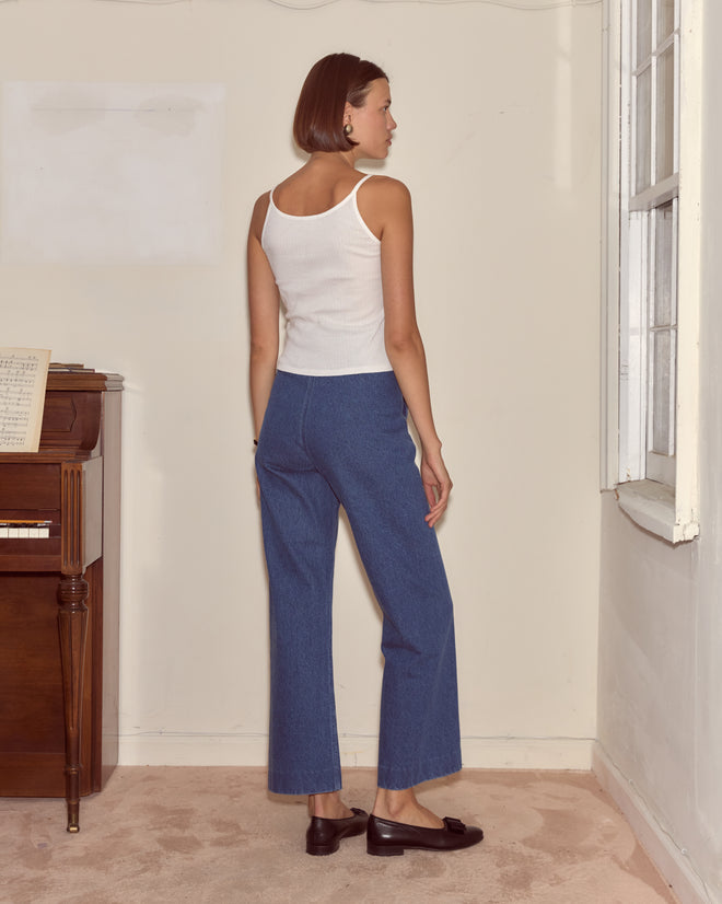 A woman stands facing away in a room with a piano and window, wearing the Companion Tank and blue jeans, creating a vintage mood.