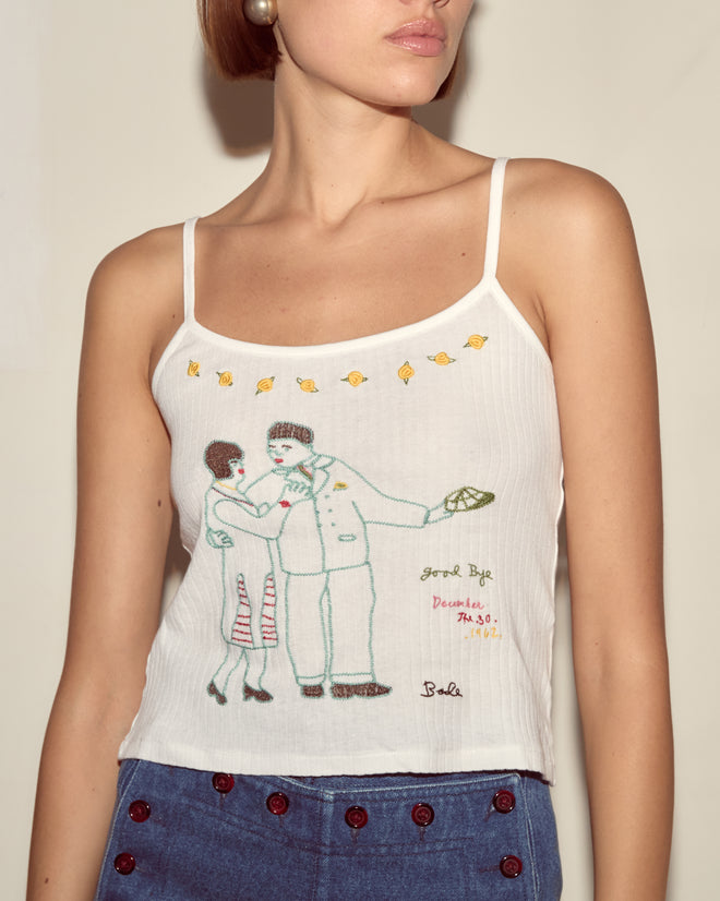 Woman wearing the Companion Tank, featuring an embroidered vintage motif with a colorful couple and goldfish, paired with blue pants—a whimsical look inspired by Moose Charlap's style.