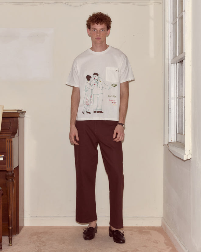 A man in a cream Companion Tee and dark pants stands indoors next to a window and a piano.