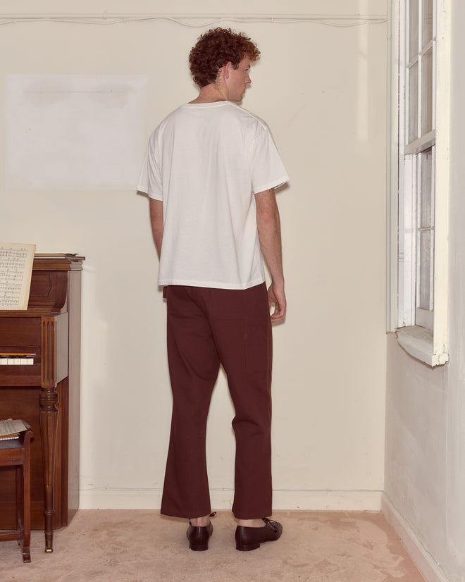 A man with curly hair stands indoors facing the wall, wearing the Companion Tee - Cream, which features a subtle vintage couple motif, paired with brown pants.