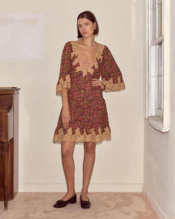 A woman in the Confetti Floral Dress with a delicate floral pattern and black flats stands indoors near a window, one hand on her hip.