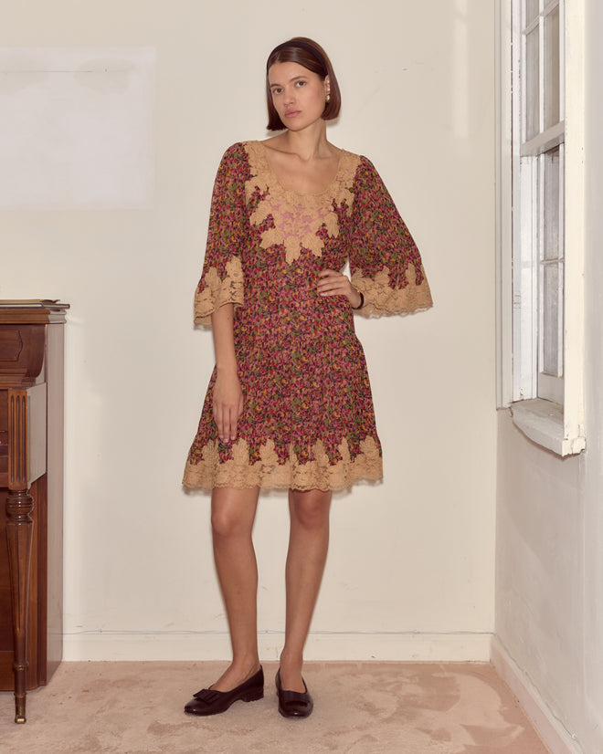 A woman in the Confetti Floral Dress with a delicate floral pattern and black flats stands indoors near a window, one hand on her hip.