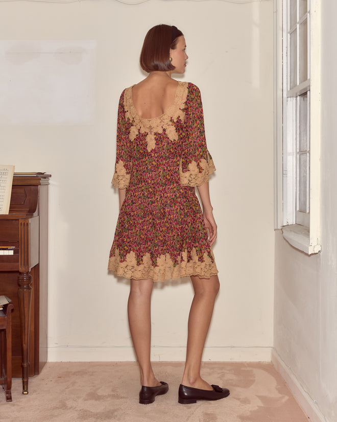 A woman in the Confetti Floral Dress, featuring silk chiffon with a delicate floral pattern and lace trim, stands by a piano and window, facing away.