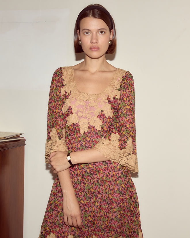 A woman wearing the Confetti Floral Dress—a silk chiffon piece with a delicate floral print—stands indoors against a plain wall, gazing into the camera.