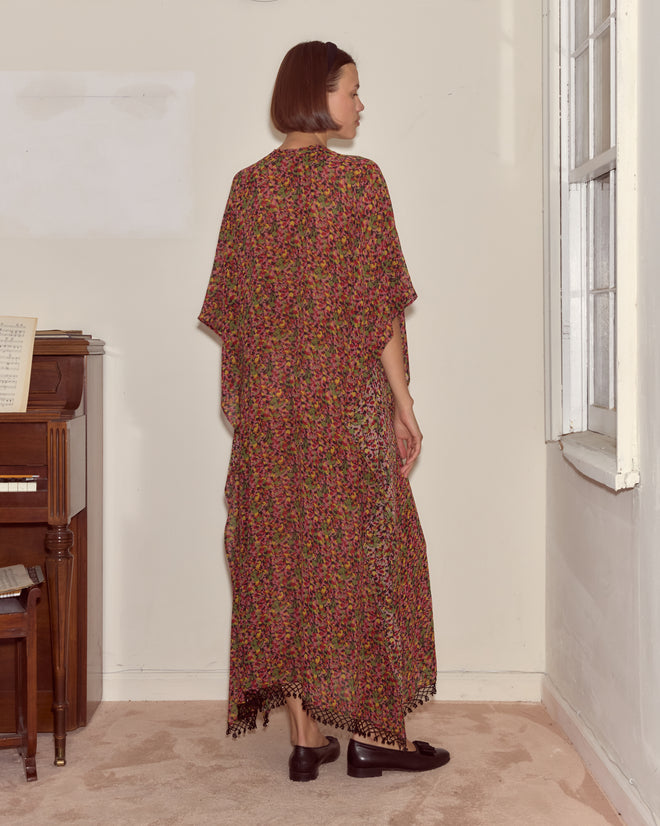 Woman in the Confetti Floral Kaftan with beaded tassel details stands by a piano, facing away by a window in a carpeted room.