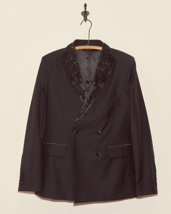 The Beaded Connaught Tuxedo Jacket, a black wool double-breasted blazer with beaded lapels, hangs elegantly on a decorative hanger against a beige wall.