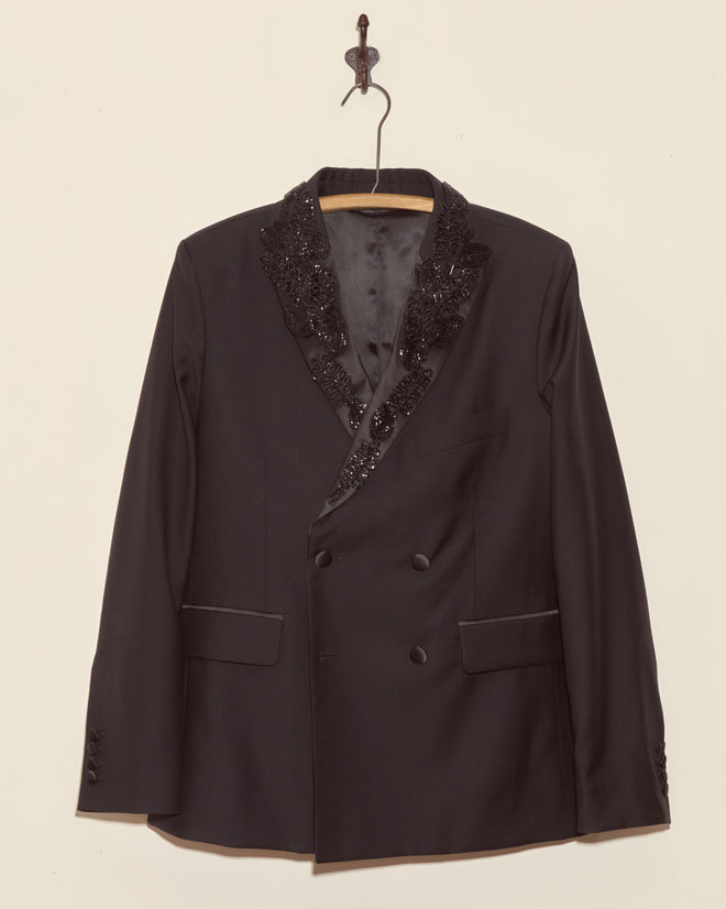 The Beaded Connaught Tuxedo Jacket, a black wool double-breasted blazer with beaded lapels, hangs elegantly on a decorative hanger against a beige wall.