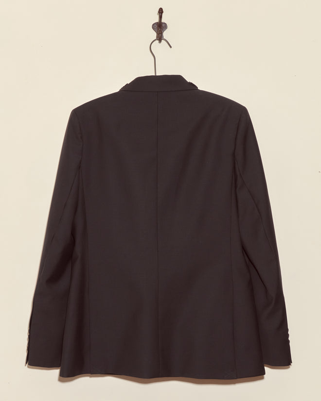 The Beaded Connaught Tuxedo Jacket, a black wool double-breasted blazer, hangs on a hook against a beige wall, displayed from the back.