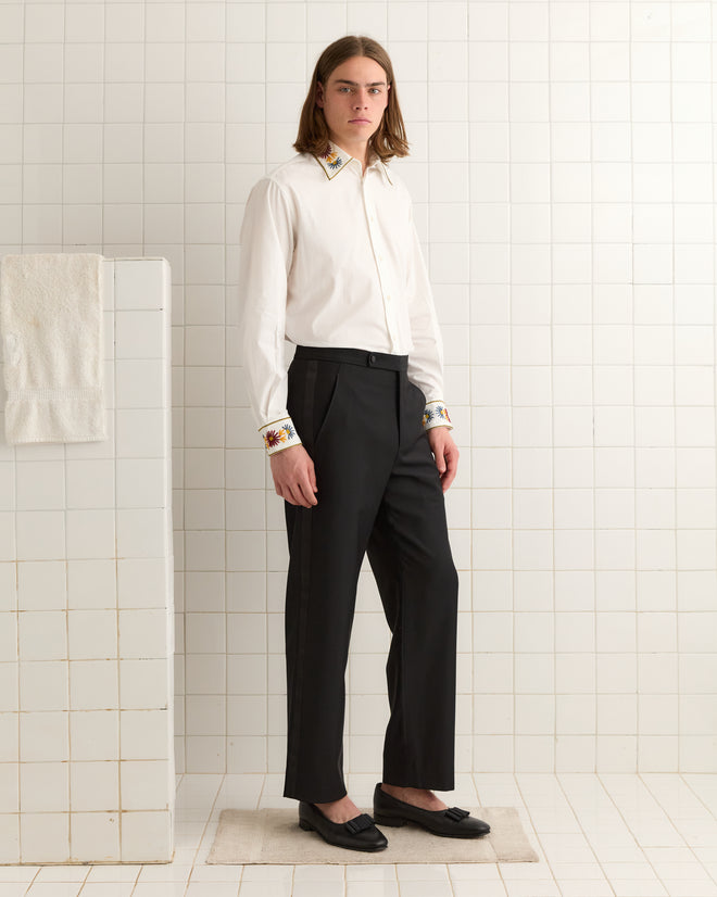 A person wearing Connaught Smoking Trousers and a white shirt stands on a tiled floor, posing in a minimalistic setting.