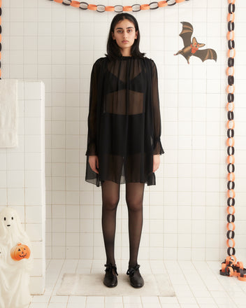 A person stands against a plain wall, wearing the Connie Dress - Black, a sheer black dress with ruffle detail over dark undergarments, paired with black tights and black shoes. Their right arm rests on the wall while their left arm hangs by their side.