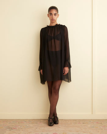 A person stands against a plain wall, wearing the Connie Dress - Black, a sheer black dress with ruffle detail over dark undergarments, paired with black tights and black shoes. Their right arm rests on the wall while their left arm hangs by their side.