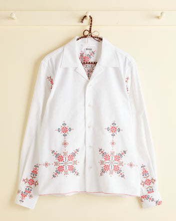 The Coral Peony Long Sleeve Shirt - S, featuring pink and red floral embroidery on the front, cuffs, and lower hem reminiscent of a 1940s cotton tablecloth, hangs on a cream-colored wall.