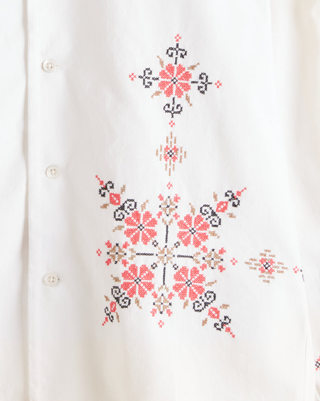 The Coral Peony Long Sleeve Shirt - S, featuring pink and red floral embroidery on the front, cuffs, and lower hem reminiscent of a 1940s cotton tablecloth, hangs on a cream-colored wall.