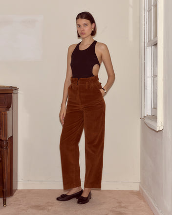 A woman stands indoors wearing a black sleeveless top, brown Corduroy Shakey Trousers with a high-rise wide leg, and black shoes.