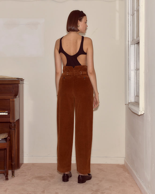 A woman stands in a room wearing the Corduroy Shakey Trousers in brown, featuring a high-rise and wide-leg design.