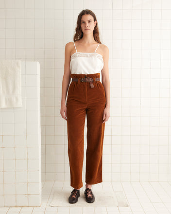 A woman stands in a tiled room wearing a white spaghetti strap top, brown Corduroy Shakey Trousers with a belt, and black shoes. A white towel is draped over a partition next to her.