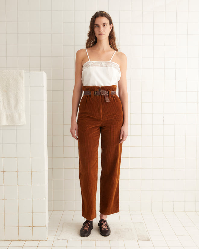 A woman stands in a tiled room wearing a white spaghetti strap top, brown Corduroy Shakey Trousers with a belt, and black shoes. A white towel is draped over a partition next to her.