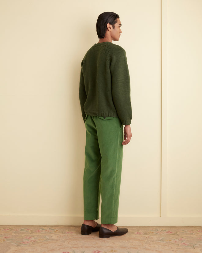 Person stands in a room, facing away, wearing a green sweater, Corduroy Side-Buckle Trousers in green with a straight-leg silhouette, and brown shoes.