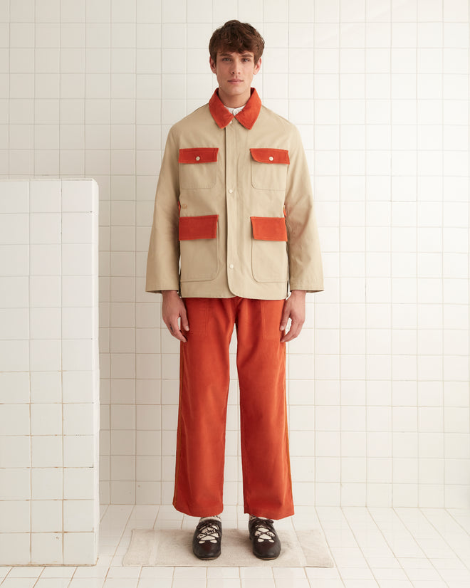 A person dressed in a beige Corduroy Trim Hunting Jacket with orange accents and matching orange pants stands in a tiled room. The ensemble, crafted from cotton twill, merges timeless style with contemporary flair.