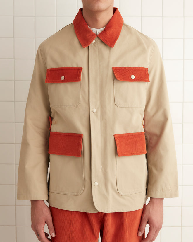 A person dressed in a beige Corduroy Trim Hunting Jacket with orange accents and matching orange pants stands in a tiled room. The ensemble, crafted from cotton twill, merges timeless style with contemporary flair.