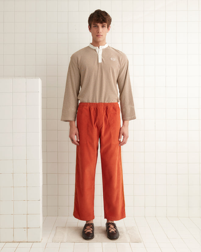 A person stands against a tiled wall, dressed in a beige striped shirt and vibrant orange Corduroy Trim Track Pants.