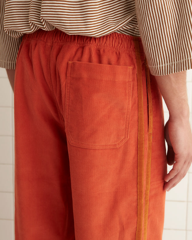 A person stands against a tiled wall, dressed in a beige striped shirt and vibrant orange Corduroy Trim Track Pants.