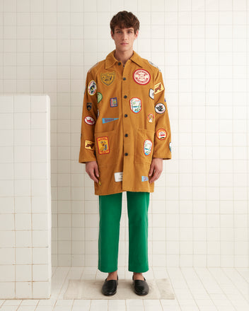 A person stands against a tiled wall, wearing the Corn Multi Patch Coat, complemented by green pants and black shoes.