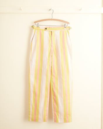 The Cotton Candy Trousers - 32, featuring a unique design with vertical yellow, pink, and orange stripes on beige deadstock material, hang on a wooden hanger against a cream wall. These one-of-a-kind pants are equipped with side-buckle waist adjusters for a perfect fit.