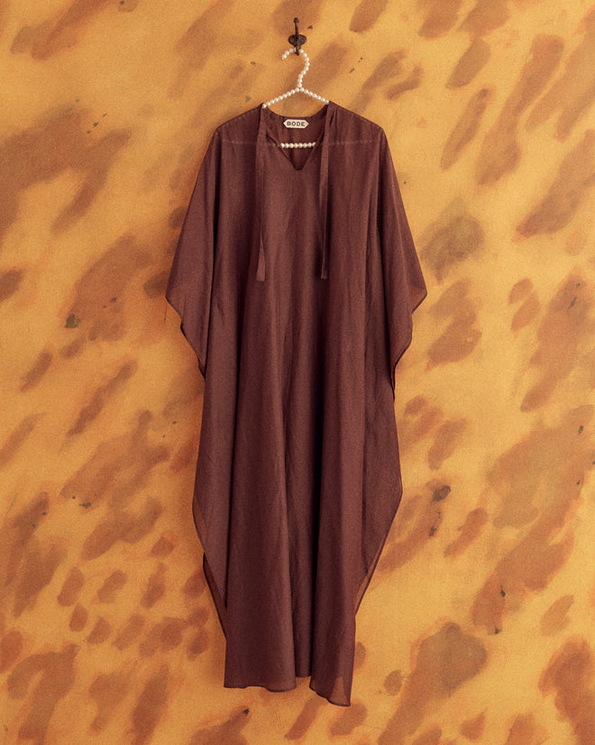 A person with long dark hair stands barefoot wearing the Cove Kaftan, which has a 1970s-inspired design, against a yellow and brown textured background.