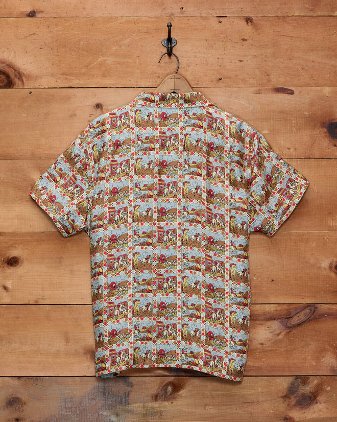 A Cowboy Comic Short Sleeve Shirt with a patterned silk design hangs on a hook against a wooden plank wall, evoking the style of classic Western comics.