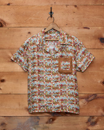 Cowboy Comic Short Sleeve Shirt displayed on a hanger against a wooden wall, featuring a brown-trimmed chest pocket for a stylish nod to classic Western comics.