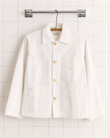 The Craft Jacket - White is a canvas workwear jacket featuring large front pockets and subtle corduroy trim, displayed on a hanger against a white tiled wall.