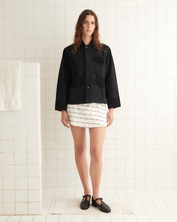 A woman wears the Craft Jacket - Black, a heavyweight cotton twill jacket with corduroy trim, paired with a white striped skirt, standing in a tiled room with white walls and floors.