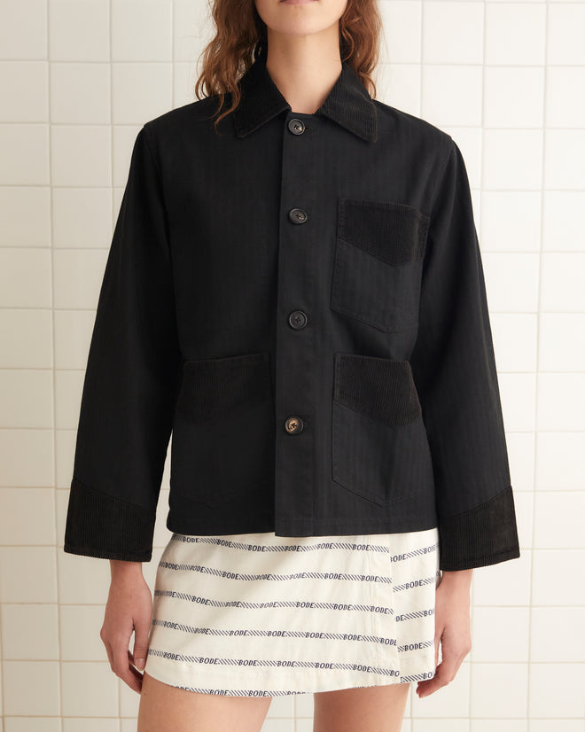 A person stands in a tiled room wearing a black button-up shirt, a white skirt with horizontal stripes, and black shoes. They have long wavy hair and are looking directly at the camera, evoking the timeless charm of the Craft Jacket with its classic corduroy trim.