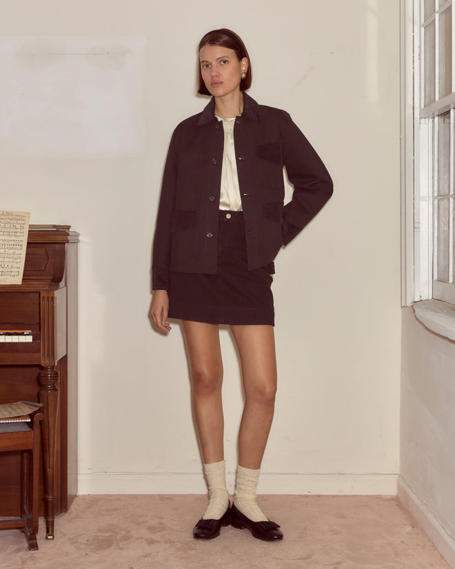 A woman stands indoors by a piano, wearing the Craft Jacket - Black, along with a skirt, white socks, and black shoes.