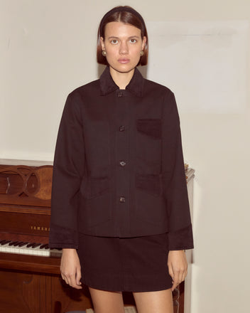 A woman in a black Craft Jacket stands in front of a piano against a plain, light-colored wall.
