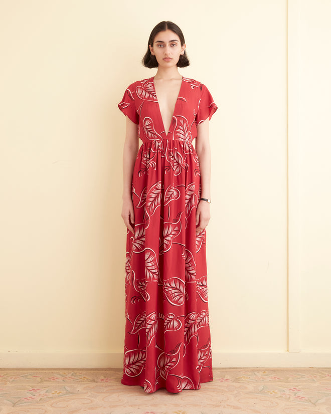 A person stands against a light yellow wall wearing the Creeping Begonia Dress, a floor-length red dress featuring a white botanical print and a deep V-neckline.