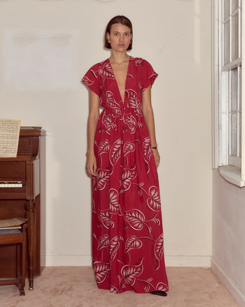 A woman stands indoors by a piano and window, wearing the Creeping Begonia Dress—a stylish red midcentury dress adorned with delicate white botanical leaf patterns.