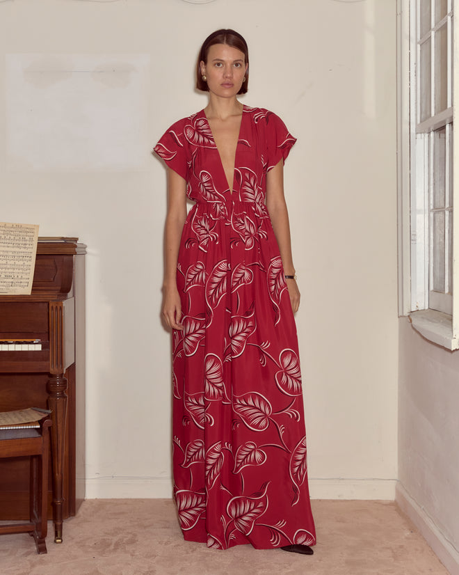 A woman stands indoors by a piano and window, wearing the Creeping Begonia Dress—a stylish red midcentury dress adorned with delicate white botanical leaf patterns.