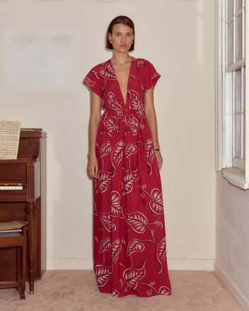 A woman in the Creeping Begonia Dress, featuring a red botanical print, stands indoors beside a piano and a window.