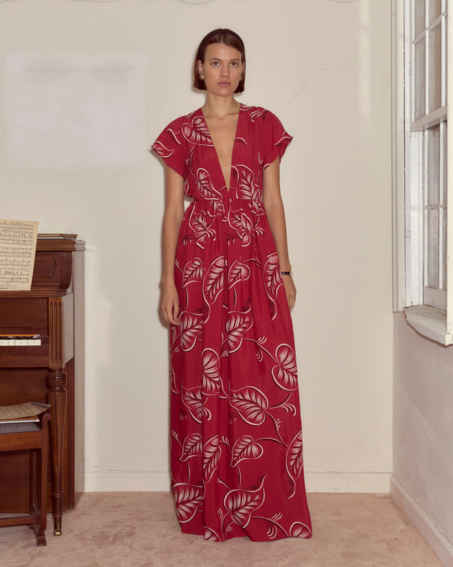 A woman in the Creeping Begonia Dress, featuring a red botanical print, stands indoors beside a piano and a window.