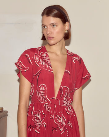 Woman wears the Creeping Begonia Dress, a deep red botanical print piece with short sleeves, looking to the side against a plain background.