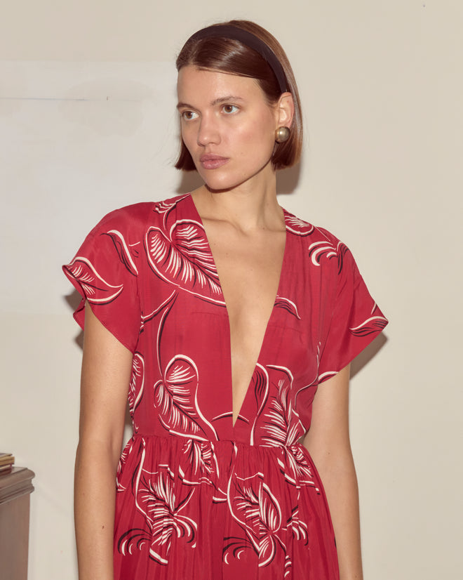 Woman wears the Creeping Begonia Dress, a deep red botanical print piece with short sleeves, looking to the side against a plain background.