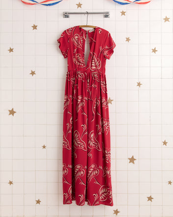 The Creeping Begonia Dress, a red maxi featuring a botanical print, hangs on a hanger against a white tiled wall adorned with gold stars for a touch of midcentury style.