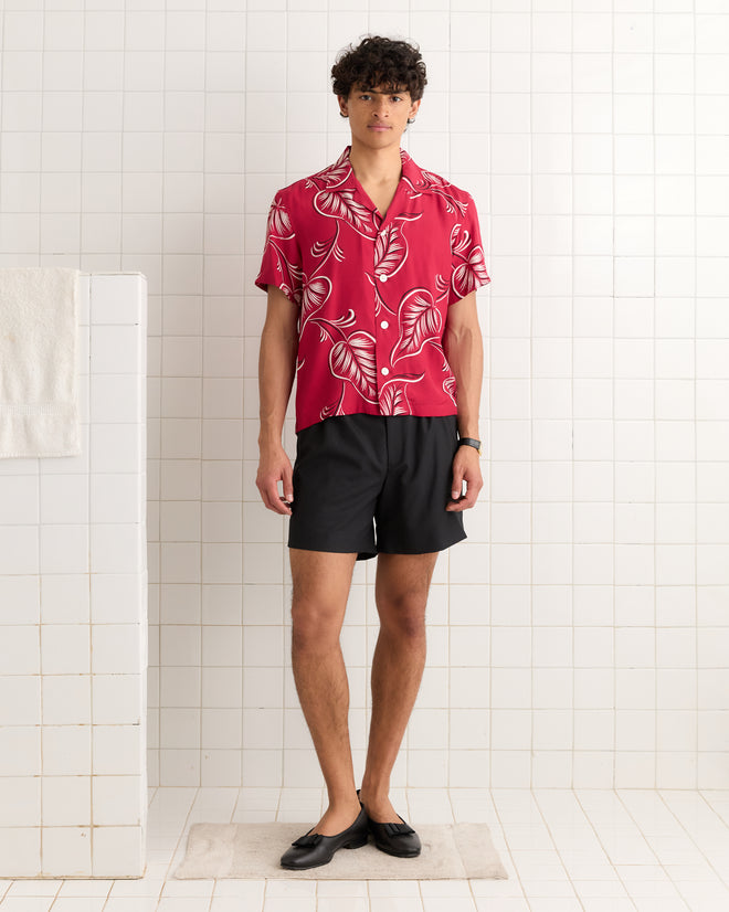 A man wears the Creeping Begonia Short Sleeve Shirt—red, oversized, with a botanical print—paired with black shorts, standing on a bath mat in a white tiled bathroom.