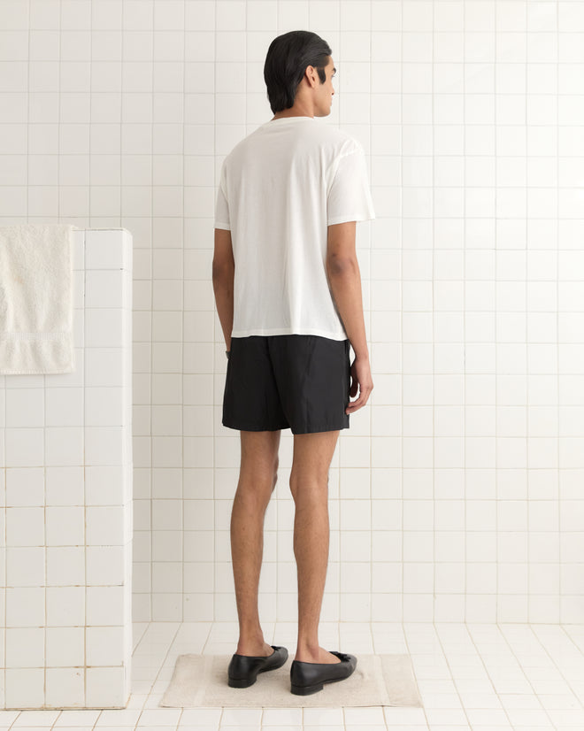 A person in a 1990s-inspired Crest Boxy Tee and black shorts stands on a bath mat in a tiled room, facing away.