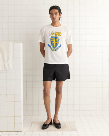 Individual in a retro-style white Crest Boxy Tee with '1852' and a crest, matched with black shorts and shoes, standing on a bathroom mat.