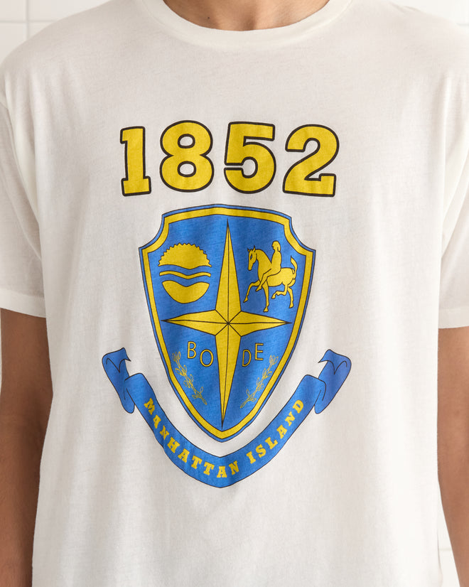 Wearing the white, lightweight cotton Crest Boxy Tee with its blue and yellow emblem, "1852," and "Manhattan Island" text, a person channels a vintage 1990s-inspired vibe.
