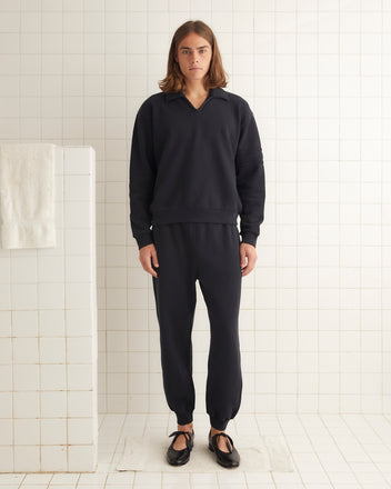 The person with long hair, dressed in a Crew Polo - Black and black jogger pants, stands facing forward in a tiled room, capturing the relaxed essence of 1980s rugby shirts.